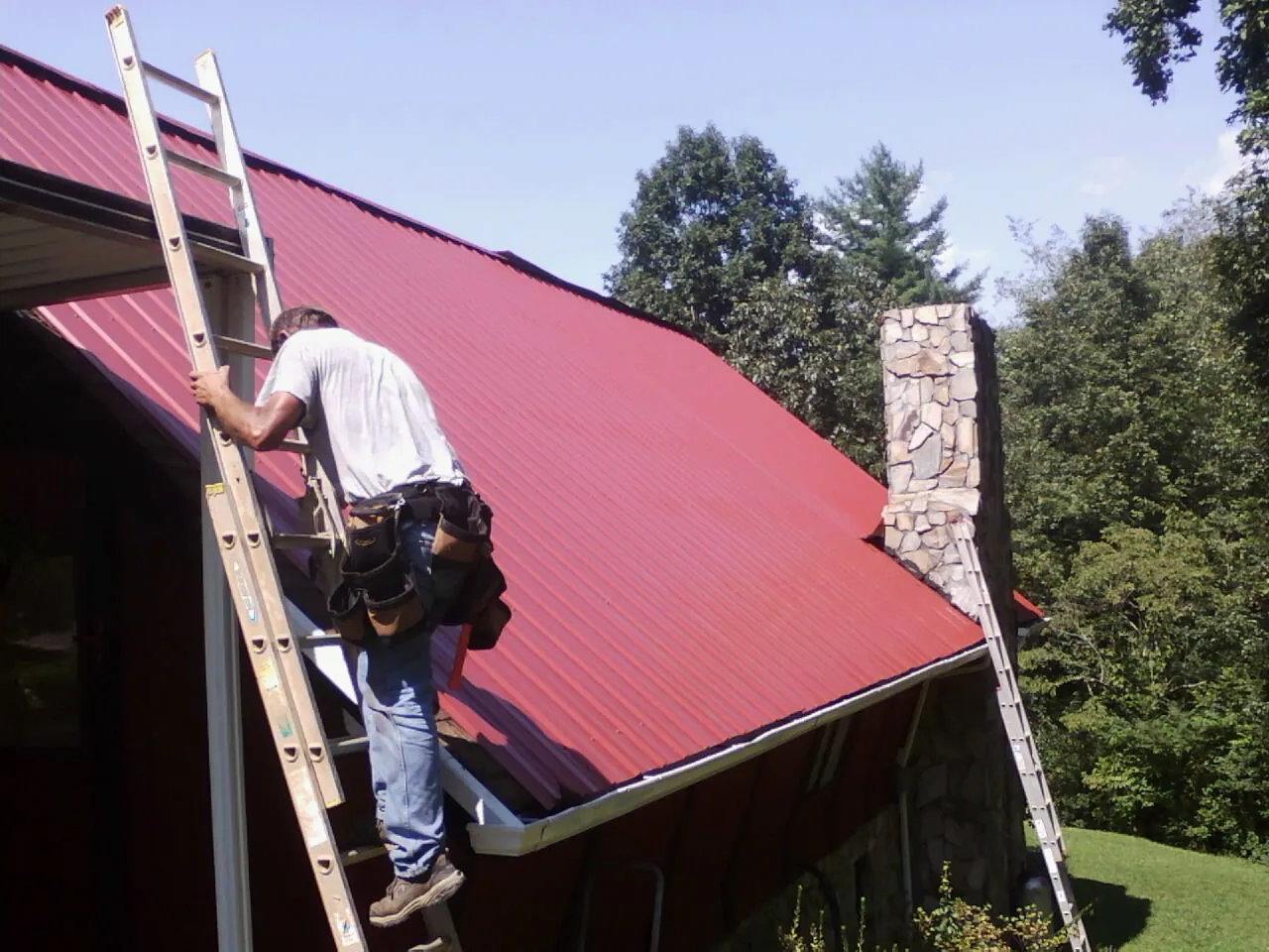Roofer on a ladder beside a red metal roof and stone chimney — Soffit & Fascia Repair by Roofing Brothers in Marbury